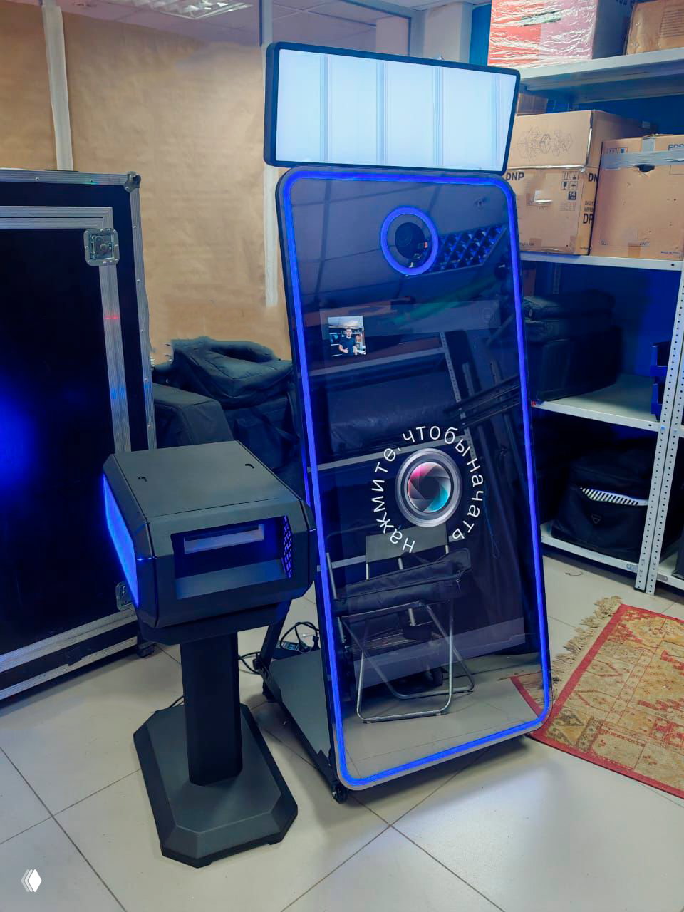 Селфи‑зеркало с RGB‑подсветкой и отдельным принтером рядом, установлено в складском/подготовительном помещении с коробками и оборудованием.