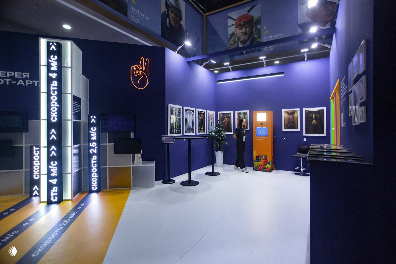 Широкий вид выставочного стенда с фиолетовыми стенами и яркой оранжевой фотостойкой у стены; видно пространство галереи и экспозицию вокруг.