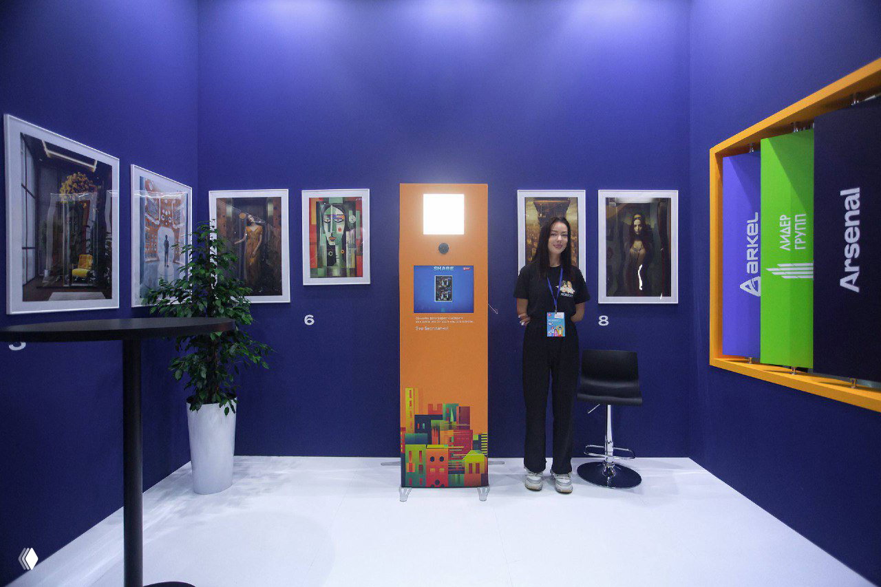 Фотостойка на белом полу выставки: сотрудница рядом, вокруг висят оформленные фотоработы и элементы оформления стенда, демонстрация размещения сервиса.