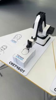 Крупный план робота‑манипулятора, рисующего портрет на бумаге; виден штриховый рисунок и подставка с надписью «СКЕТЧБОТ», акцент на процессе механического рисования.