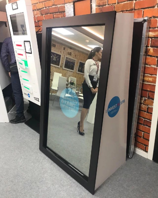 Стационарное зеркальное устройство для фотозоны на выставочном стенде у кирпичной стены; видна отражённая сотрудница и часть оборудования.