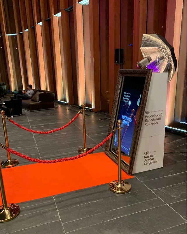 Селфи‑зеркало в стойке при входе на мероприятие: красная дорожка, стойки-ограждения и подсвеченные колонны в фойе.