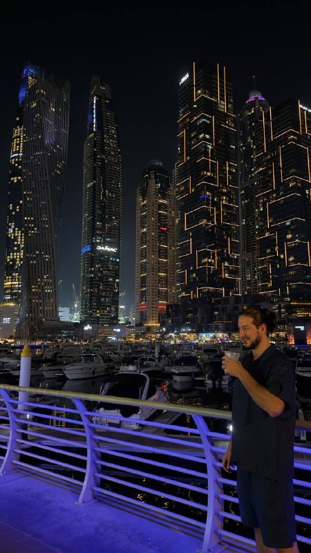Ночное фото на набережной Dubai Marina: освещённые небоскрёбы, причалы с яхтами и мужчина в тёмной одежде на переднем плане.