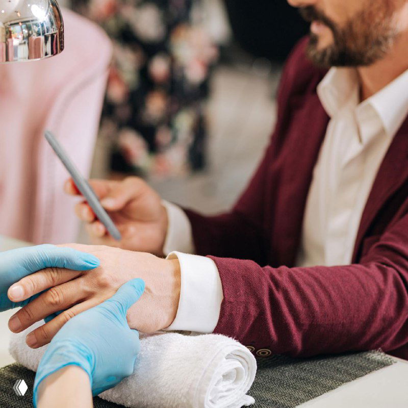 Мужчина в пиджаке получает маникюр: мастер в перчатках держит руку клиента, видны салонная обстановка и деловой образ.