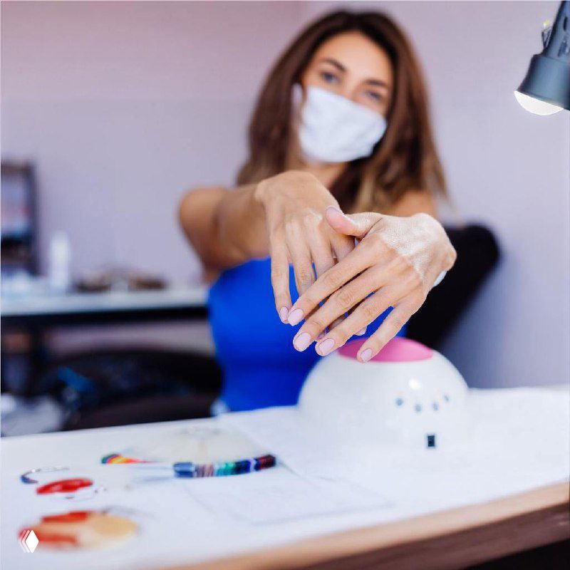 Клиентка показывает руки с готовым маникюром у лампы для сушки; на фоне рабочий стол мастера с инструментами и материалами для покрытия.