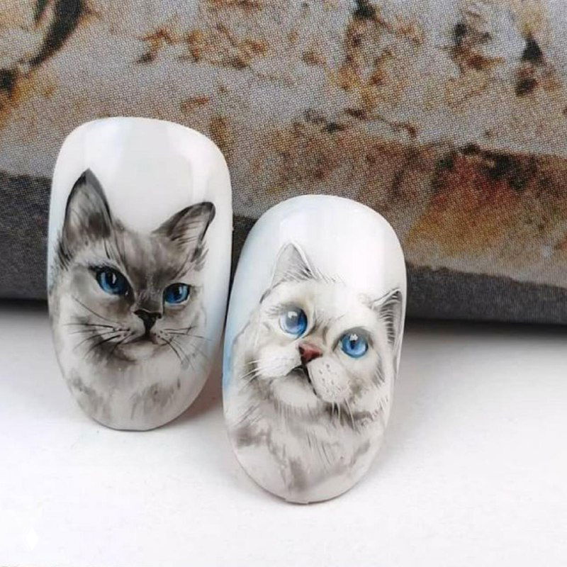 Два образца ногтей с фотореалистичным рисунком кошки на белой базе: детальная мордочка с голубыми глазами, пример авторской росписи акварелью в маникюре.