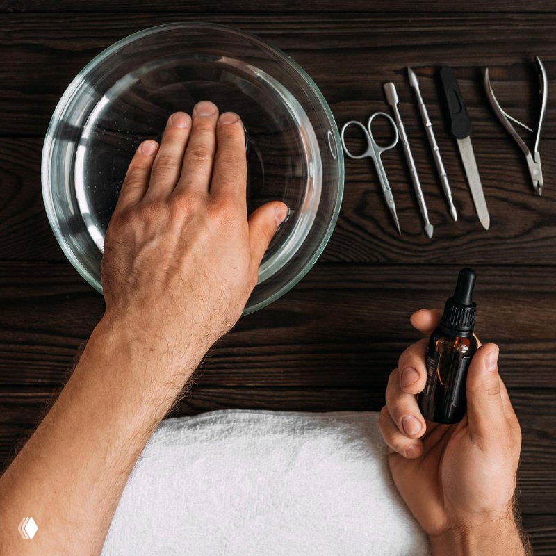 Рука мужчины над миской с водой и набор инструментов для маникюра: щипчики, ножницы и масло для кутикулы на тёмном фоне.