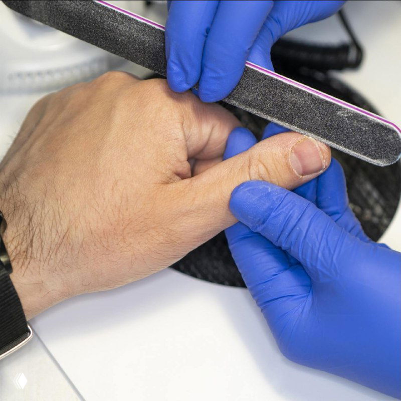 Процесс: мастер в синих перчатках подпиливает мужской ноготь пилкой, крупный план пальцев и аккуратная рабочая поверхность.