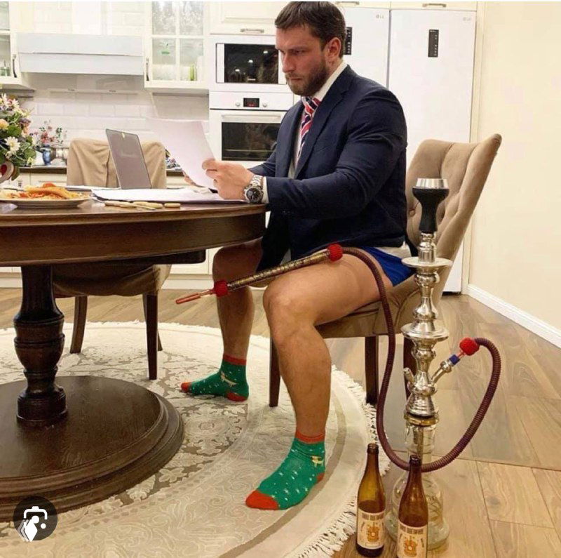 Мужчина в пиджаке и галстуке без брюк сидит за столом с кальяном и бутылками, сочетая формальный верх и домашний низ в кадре.