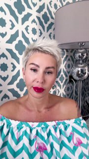 Портрет женщины с короткими светлыми волосами и яркой красной помадой на фоне узорчатой стены и настольной лампы.