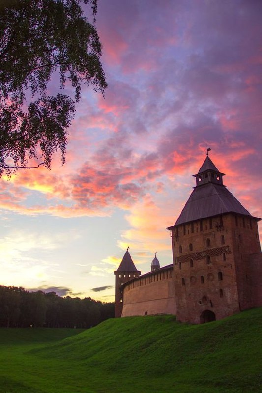Древний кремль на холме при закате, зелёные луга вокруг и драматичное розовое небо над историческим Великим Новгородом
