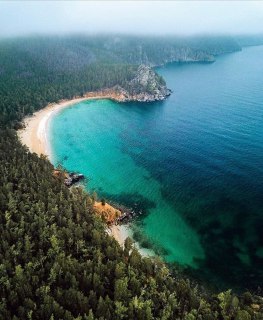 Песчаный пляж и изумрудные воды озера Байкал с высокой скалистой береговой линией и зелёной прибрежной растительностью