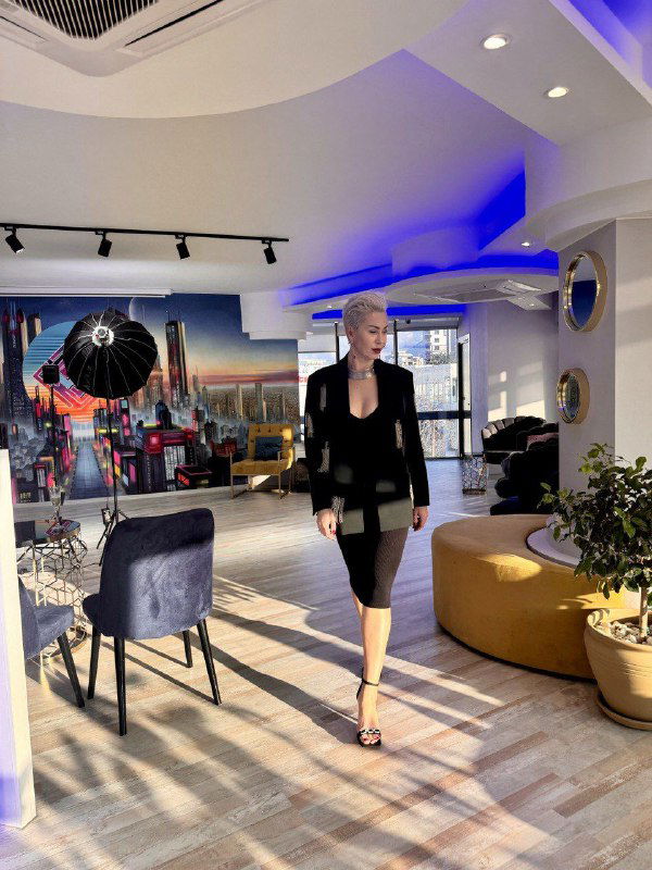 Женщина в строгом платье проходит по интерьеру студии с современным дизайном, фототехникой и яркими декоративными акцентами.