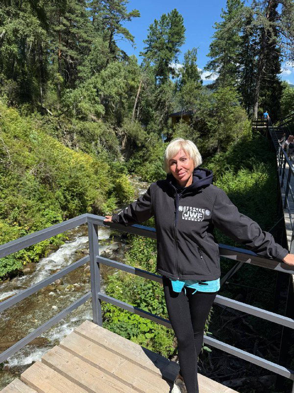 Наталья стоит на металлическом мостике в лесном ущелье; на фоне горный поток, хвойные деревья и ясное летнее небо, автор в фирменной куртке блога.