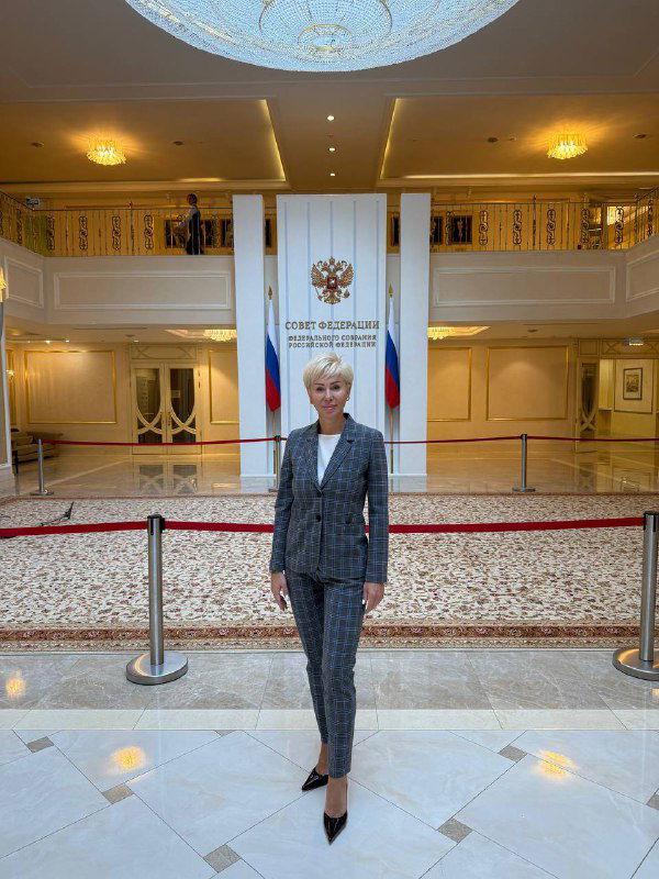 Наталья Будько стоит в холле Совета Федерации перед государственным гербом и флагами, полный рост в деловом костюме.