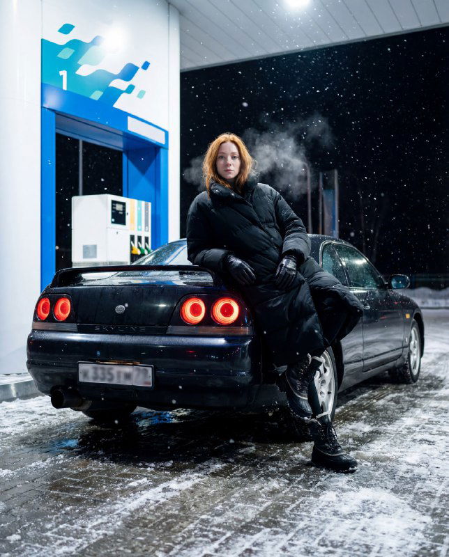 Женщина в тёмном пальто опирается на багажник купе у заправки ночью: видны снежные хлопья, пар дыхания и мокрый отражающий асфальт.