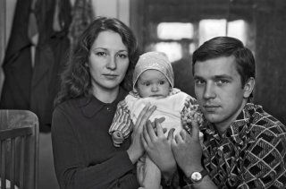 Чёрно‑белая семейная фотография: мать, отец и младенец на руках у родителей в интерьерной сцене, выражения лиц спокойные, взгляд в камеру