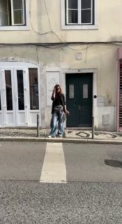На фото женщина в повседневной одежде стоит на пешеходном переходе перед фасадом дома с зелёной дверью; уличный дневной кадр.