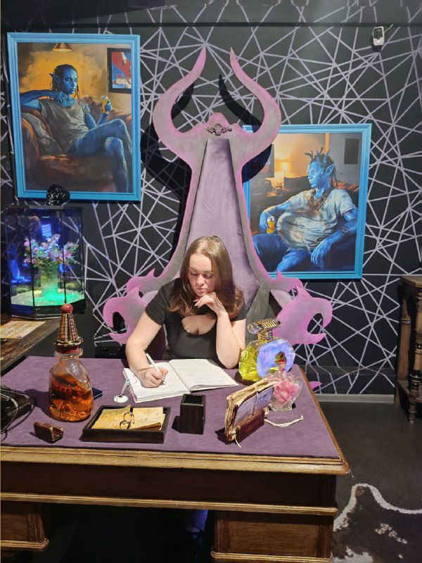 Автор сидит за стилизованным столом в игровой комнате квеста среди декораций, бок о бок с реквизитом и театральной обстановкой для фото.