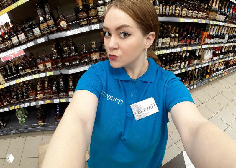 Селфи женщины в синей форме на фоне стеллажей с алкоголем в супермаркете: витрины и бутылки создают контекст рабочего места.