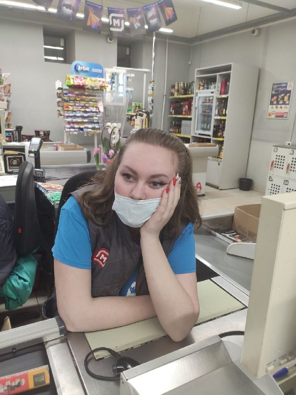 Женщина в форме продавца сидит за кассой с маской на подбородке, выглядит уставшей: коробки и полки магазина на заднем плане.