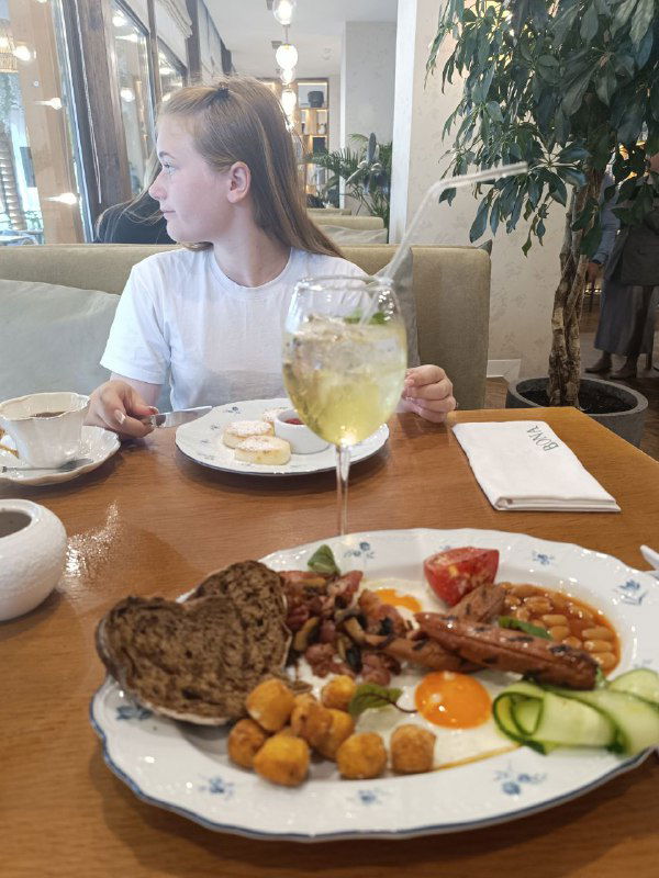 Завтрак в ресторане: тарелка с яйцами, хлебом и овощами на переднем плане, за столом сидит девушка в белой футболке, тихая утренняя сцена.