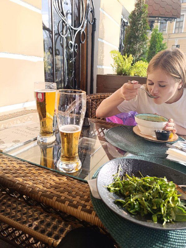 Уличный стол на летней веранде: два бокала пива и миски с едой, девочка за столом ест суп — лёгкая атмосферная сцена городского отдыха.