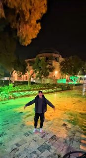 Человек стоит ночью на влажной каменной площади перед куполообразным зданием; зелёная подсветка и деревья создают атмосферу паломничества и исследования.