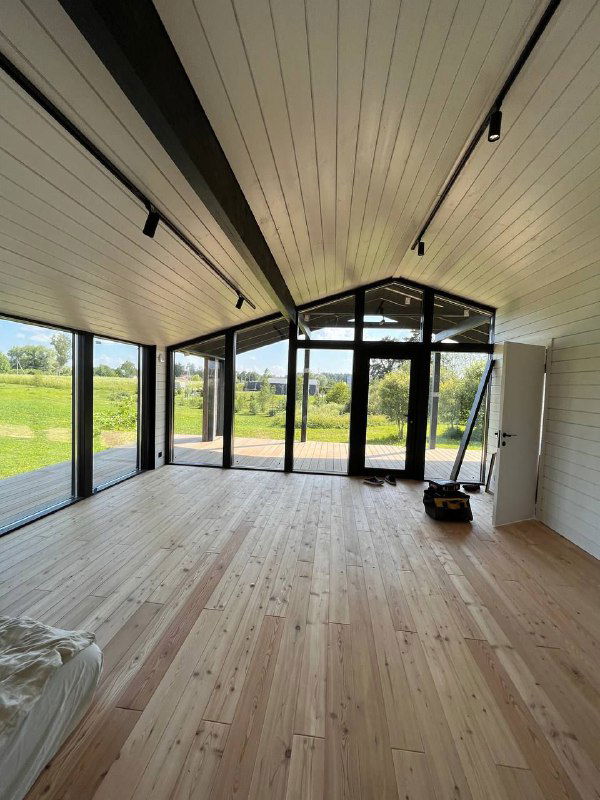 Внутренний вид барнхауса с деревянным полом, белым потолком и большими панорамными окнами; вид на зелёный участок за домом.