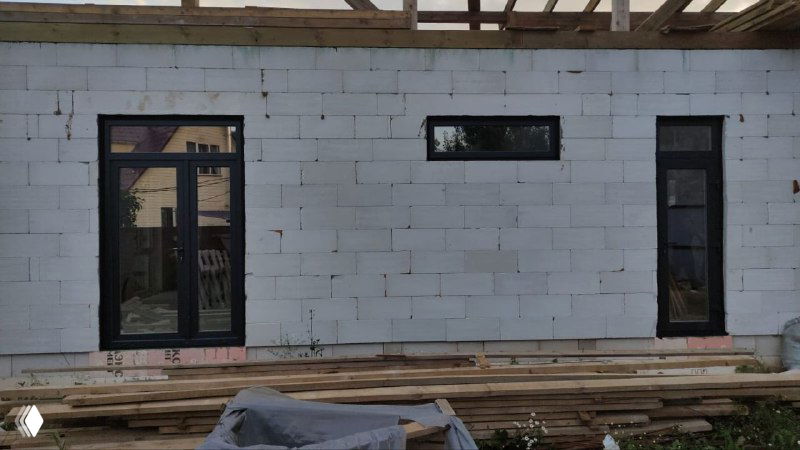 Длинная стена из газобетонных блоков с несколькими стеклянными проёмами и установленными тёмными рамами в антрацитовой ламинации.