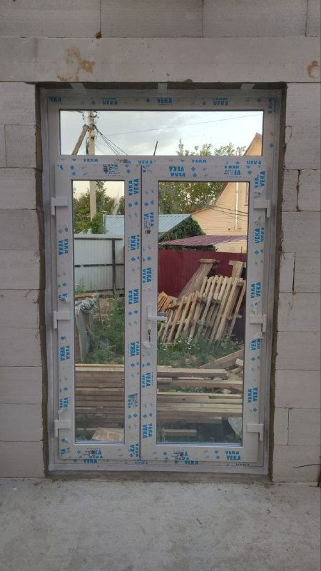 Окно крупным планом с транспортной защитной плёнкой и видимыми строительными материалами за проёмом, этап внутренней отделки ещё не завершён.