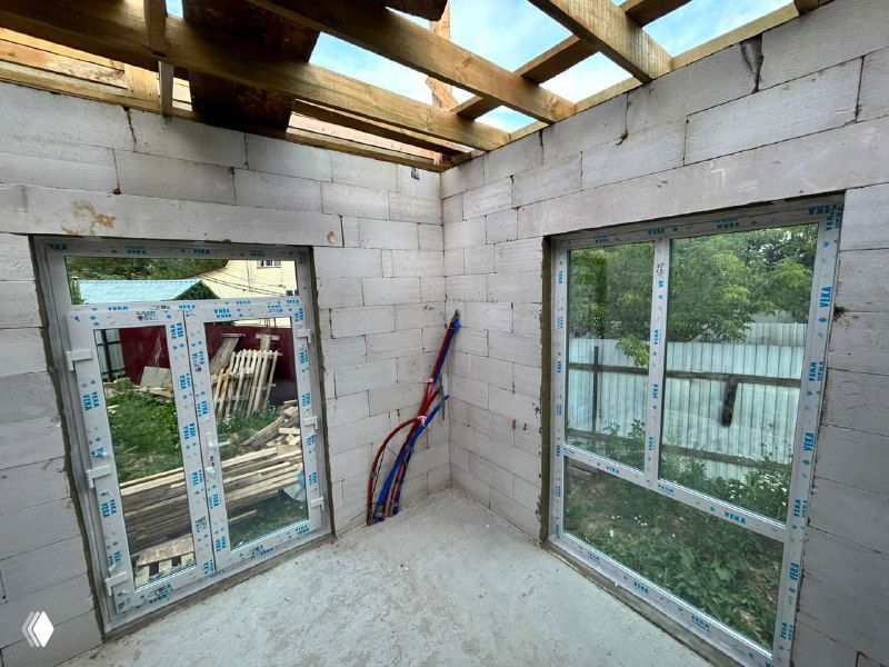 Интерьер: два больших оконных проёма на одном уровне, вид на сад через установленные стеклопакеты, ещё заметны коммуникации и рабочие инструменты.