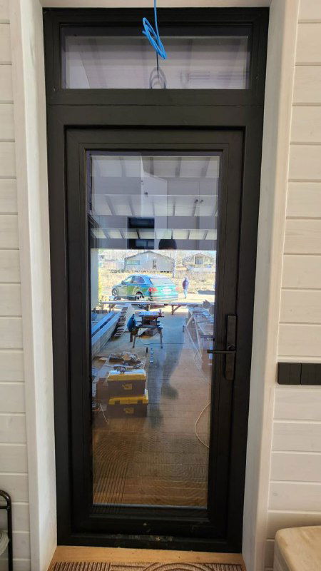 Входная дверь в черной рамке Gealan S9000 82 мм на фоне мастерской, видно оборудование, упаковку и отражения в стекле