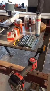 Рабочий стол в оконной мастерской с деталями для монтажа: профили, баллоны монтажной пены, направляющие, метизы и рулетка на фоне инструмента.