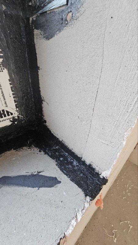 Крупный план внутренней стороны откоса с нанесенным черным герметиком по линии примыкания рамы и подготовленными слоями для внешней защиты.