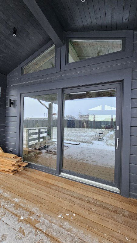 Раздвижная система в закрытом положении на террасе: тёмная рама, большие стеклопакеты и вид на внутреннюю часть дома через стекло.