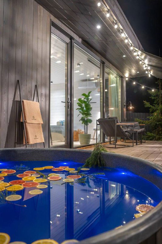 Ночной кадр террасы с гирляндами и столовой зоной рядом с панорамными окнами, мягкое декоративное освещение внутри и снаружи дома.