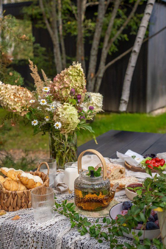 Снимок пикника на свежем воздухе рядом с домом: стол, плетёная корзина и букеты, подчёркивающие атмосферу уютного загородного отдыха у дома.