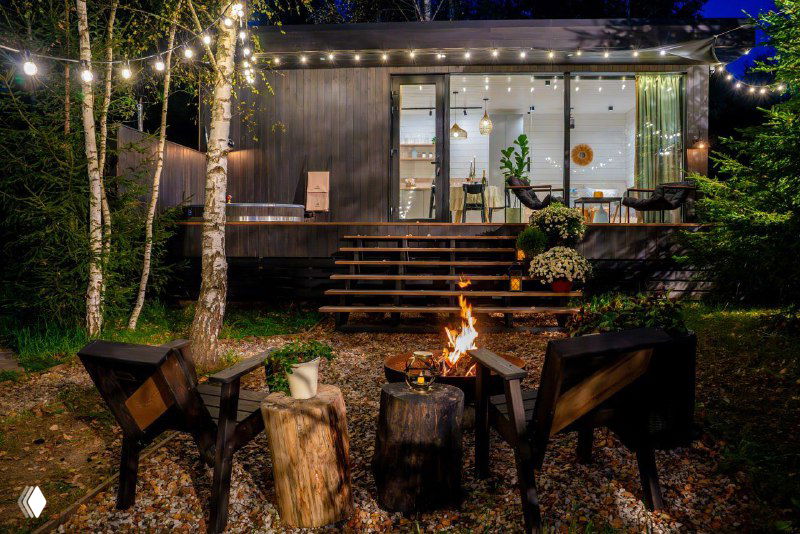 Общий вид на освещённую террасу и крыльцо дома вечером: ступени, растительность и интерьерные огни через панорамные окна.