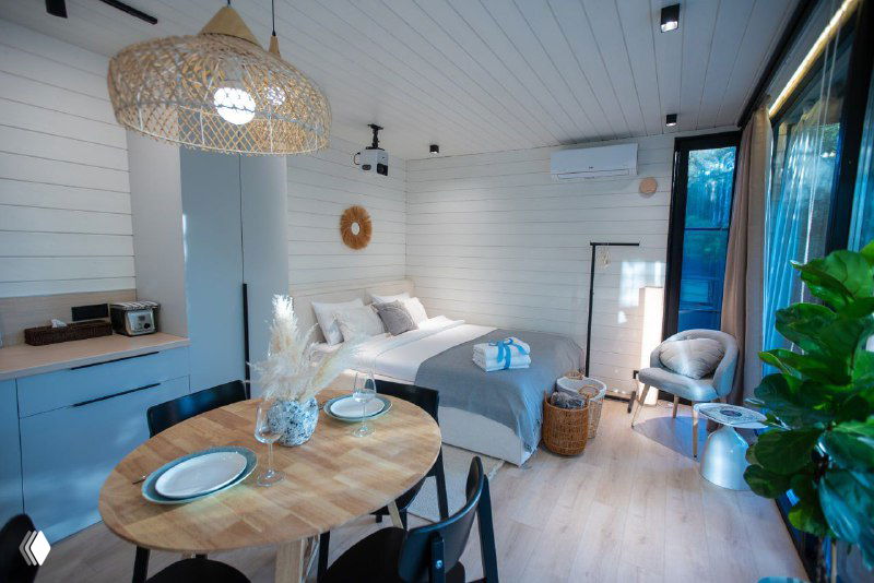 Интерьер: светлая открытая кухня-гостиная в каркасном доме с деревянной отделкой, обеденным столом и большими стеклянными окнами до пола.