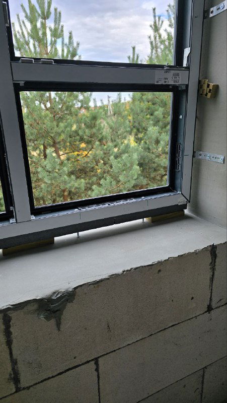 Крупный план нижней части окна: герметизация стыка, подоконник и вид на зелёную зону за окном; серый профиль и аккуратные швы монтажа.