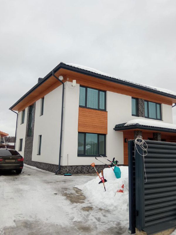 Вид готового двухэтажного дома с установленными окнами Schuco: контраст тёмных рам, декоративная облицовка фасада и снег у ворот.