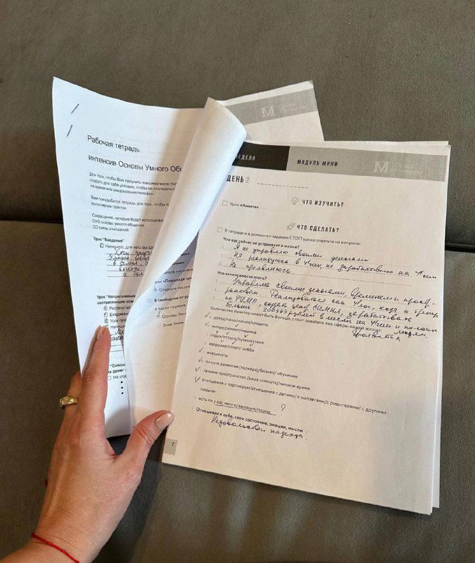Рука держит страницы учебного материала и рабочей тетради с заметками и заполненными заданиями — крупный план, процесс работы ученицы.