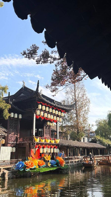 Традиционное здание у воды с яркими китайскими фонарями и осенними ветвями на фоне синего неба, вечерняя сцена на набережной у канала.