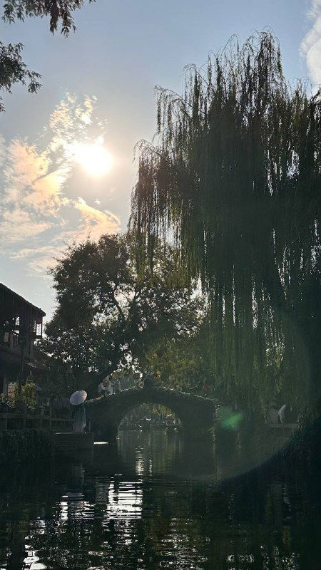 Солнце пробивается сквозь ветви ивы у канала, видна арка каменного моста и мягкие отражения деревьев на поверхности воды традиционного городка.