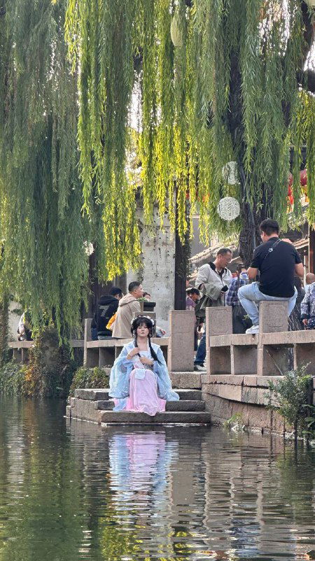 Люди на набережной у воды среди свисающих ив, молодая женщина в маске сидит у берега — городская туристическая сцена у канала в старом районе.