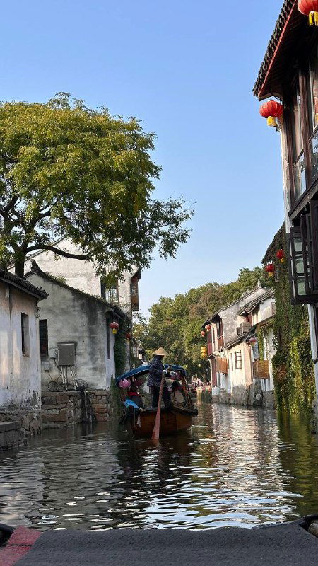 Лодка плывёт по каналу между зданиями, вдоль берега видны деревья и прохожие — традиционная водная улица в спокойном туристическом районе Китая.