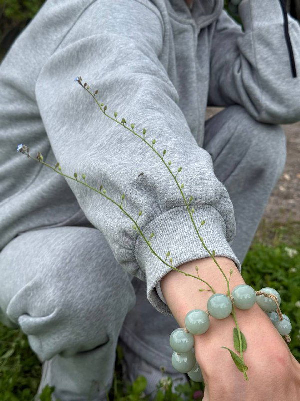 Крупный план руки в сером худи с браслетом из больших зелёных шариков и тонкими травинками — акцент на простых элементах и деталях.