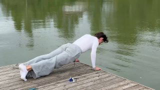 Человек в белой футболке и светлых штанах делает упор лёжа на деревянном пирсе у воды — упражнение на устойчивость и связь с природой.