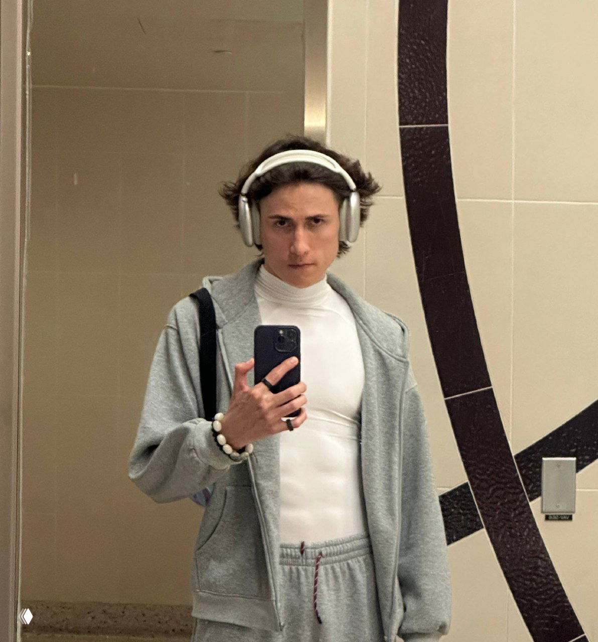 Автопортрет в зеркале: человек в светло‑сером худи и наушниках держит смартфон, отражение в ванной/раздевалке на заднем плане.